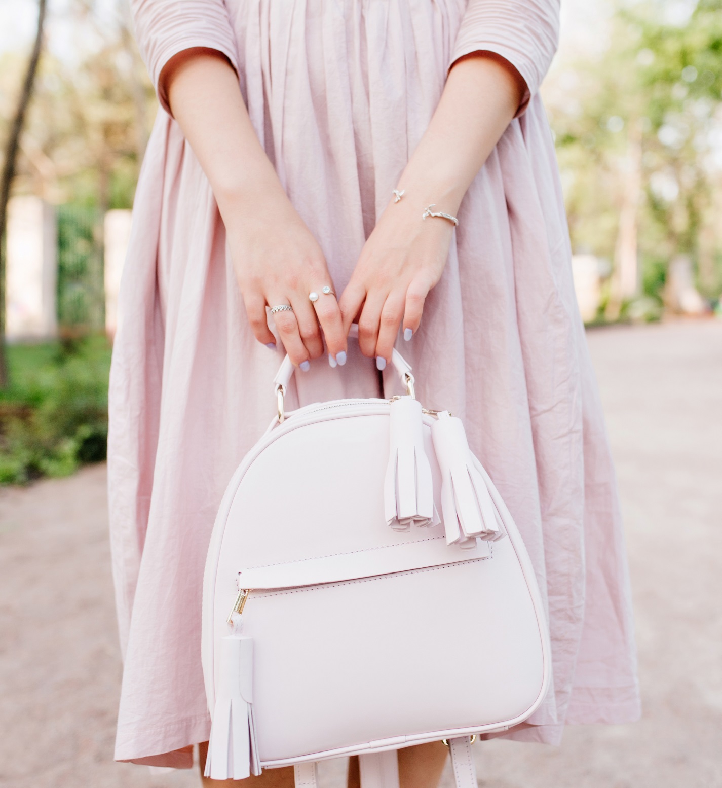 Girl with elegant manicure posing during spending time outdoor, standing on park alley. Young lady wearing rings and bracelet holding leather backpack walking outside in spring day.